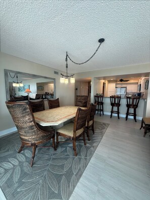 A dining area with a large table and chairs, a hanging light fixture, and a kitchen in the background.