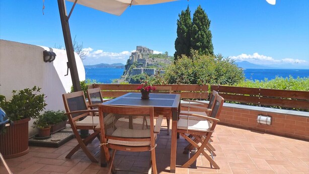 Une terrasse extérieure avec une table et des chaises, un grand parasol, et une vue sur un bâtiment historique et la mer.