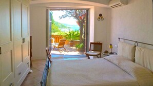 Une chambre avec un grand lit, une petite table et une chaise. Une porte coulissante donne accès à un balcon avec vue sur la mer.