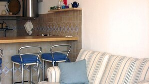 Un espace de vie compact comprenant un canapé à rayures, deux tabourets de bar, un coin cuisine avec une cuisinière et un mur recouvert de carreaux.