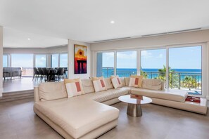 A spacious living room with a large L-shaped beige sofa, a round coffee table, and a dining area with chairs and a table. - Apartment M601 - Spectacular Beachfront Penthouse! (Scarborough)