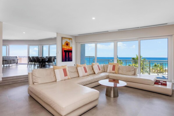 A spacious living room with a large L-shaped beige sofa, a round coffee table, and a dining area with chairs and a table.