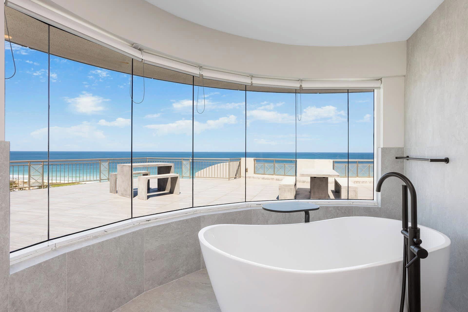 A modern bathroom with a large bathtub, a view of the ocean through floor-to-ceiling windows, and a balcony with outdoor seating.