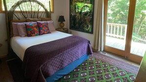 Un dormitorio con techo de madera, una cama con colcha morada, alfombra verde estampada, un ventanal con vistas a la vegetación y un cuadro en la pared.