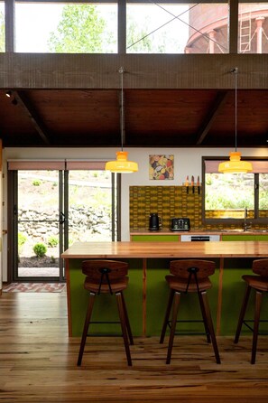 Une cuisine moderne avec un îlot central en bois, des tabourets de bar, des luminaires suspendus et un mur en pierre visible à travers la porte en verre.