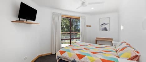 Un dormitorio con una cama, un televisor montado en la pared, un ventilador de techo, una ventana con cortinas y un cuadro enmarcado.