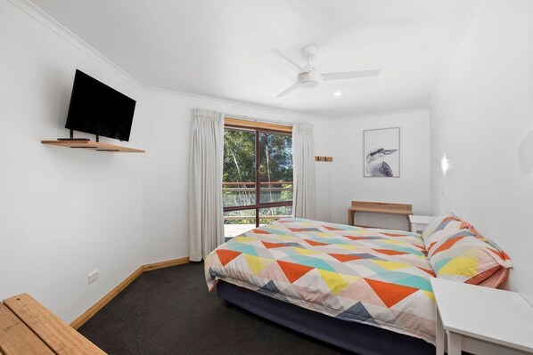 A bedroom with a bed, a TV mounted on the wall, a ceiling fan, a window with curtains, and a framed picture on the wall.