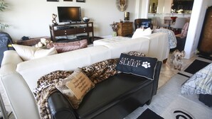 Una sala de estar con un sofá blanco, un reposapiés negro y un televisor sobre un mueble de madera. Hay almohadones decorativos y un perro en el suelo.