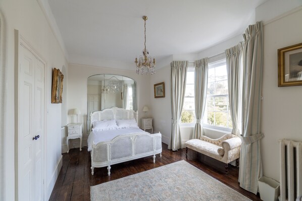 A bedroom with a four-poster bed, a sitting bench, a mirror, a chandelier, and a window with curtains.
