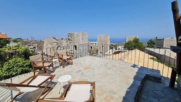A rooftop terrace with stone flooring, wooden lounge chairs, and a view of historical stone buildings and the sea. - Old Tower-House in Mani peninsula (LAGIA)