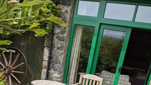 Un patio de piedra con una mesa redonda y dos sillas de madera, una rueda de madera y una puerta verde que da a una habitación con cama.