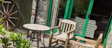 A stone patio with a round table and two wooden chairs, a wooden wheel, and a green door leading to a room with a bed.