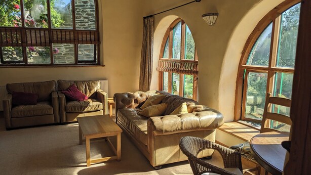 Una sala de estar acogedora con sofás de cuero, una mesa de centro de madera y un sillón de mimbre. Amplias ventanas en arco ofrecen luz natural y vistas al verdor.