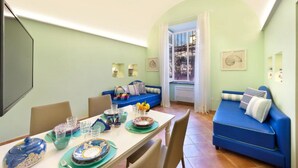 A dining area with a table set for two, a blue sofa, and a window with curtains. - Henry House (Sorrento)