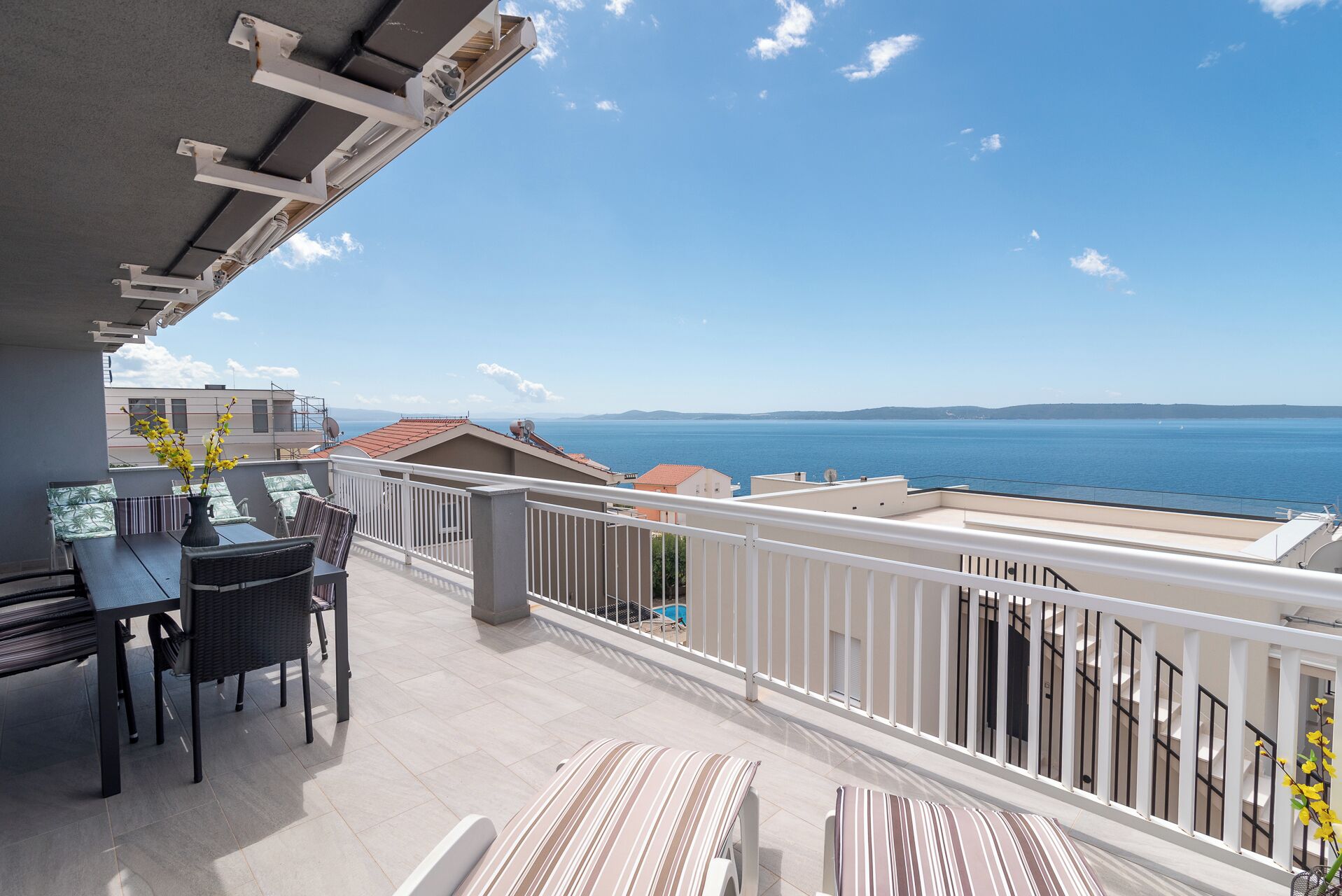 A balcony with a table and chairs, a view of the sea, and buildings in the background.