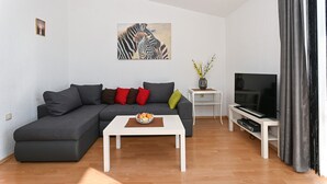 Ein modernes Wohnzimmer mit einem grauen Ecksofa, einem weißen Couchtisch, einem Flachbildfernseher und einem kleinen weißen Beistelltisch.