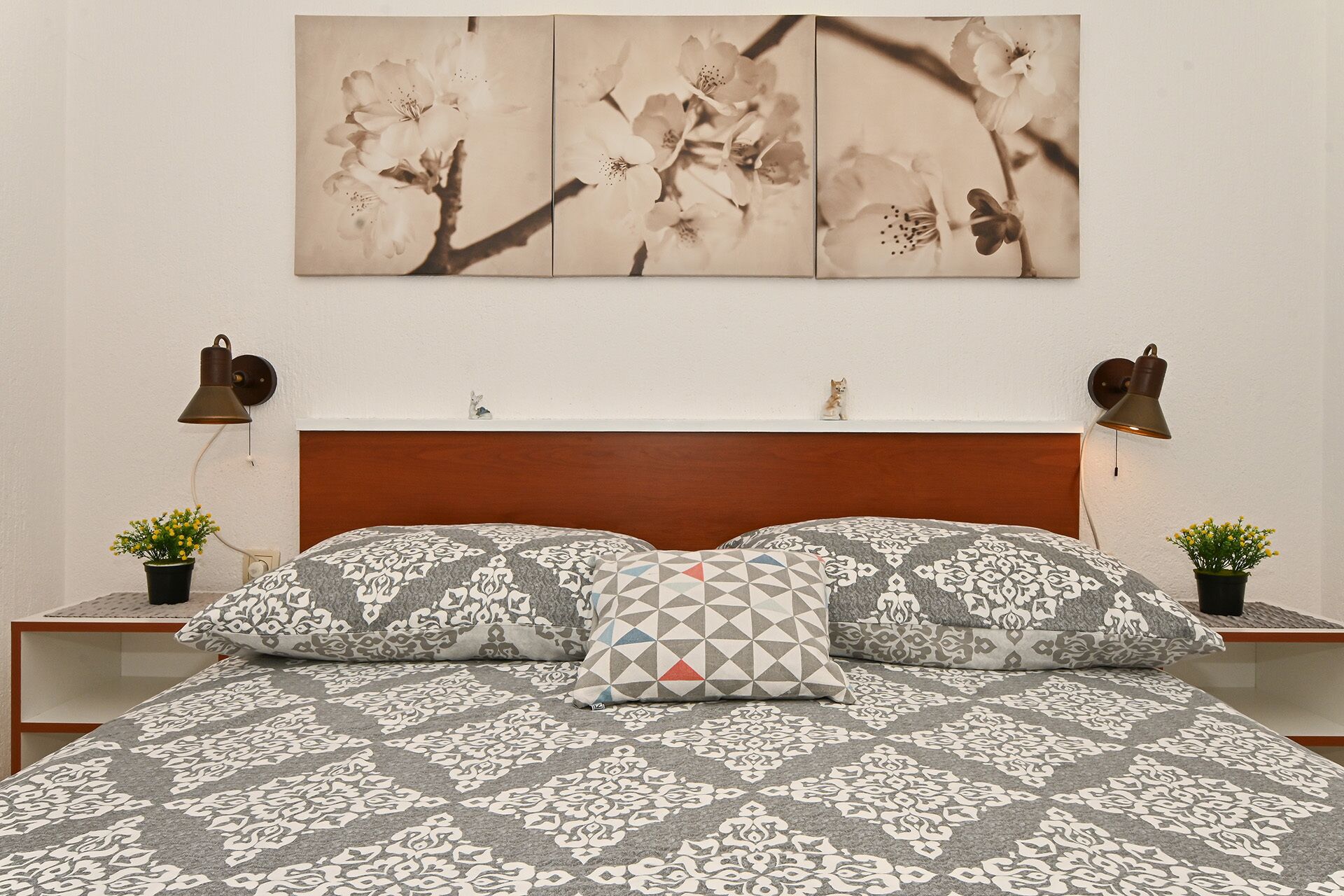 A bedroom with a patterned bedspread, two matching pillows, a wooden headboard, and wall-mounted lamps.