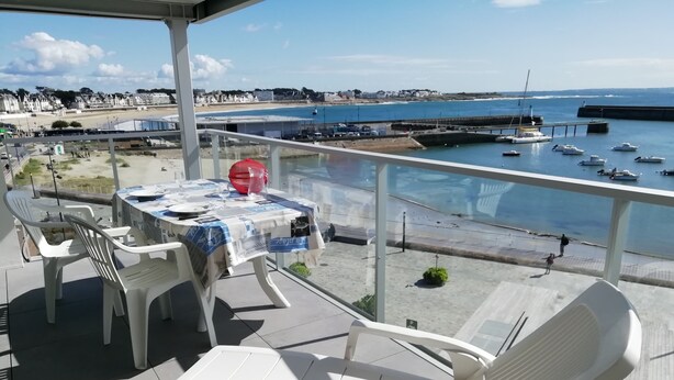 Una terraza con mesa y sillas, con vista a una playa y un puerto deportivo.