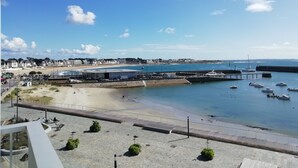 Vista costera con una playa arenosa, un paseo marítimo y un puerto deportivo con barcos.