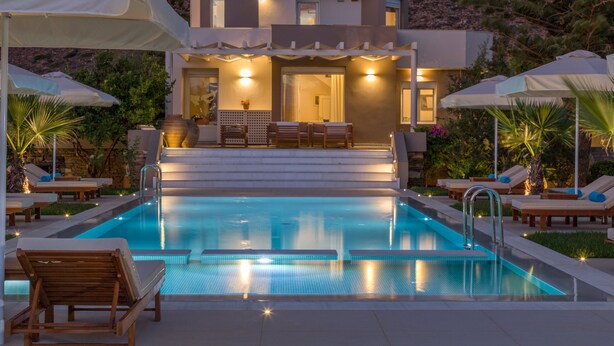 Una piscina con sillas de descanso y sombrillas, un edificio con terraza y montañas al fondo.