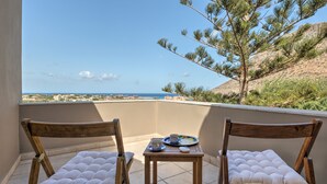 Un balcón con dos sillas de madera y una pequeña mesa, con vista a un paisaje costero que incluye el mar y edificios a lo lejos.