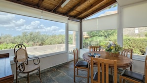 Un patio cubierto con una mesa y sillas de madera, una cesta de mimbre con flores y vistas a la vegetación y una carretera.