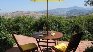 Ein runder Holztisch mit zwei Stühlen, einem gelben Sonnenschirm und einer Weinflasche auf einer Terrasse mit herrlichem Blick auf Hügel und Weinberge.