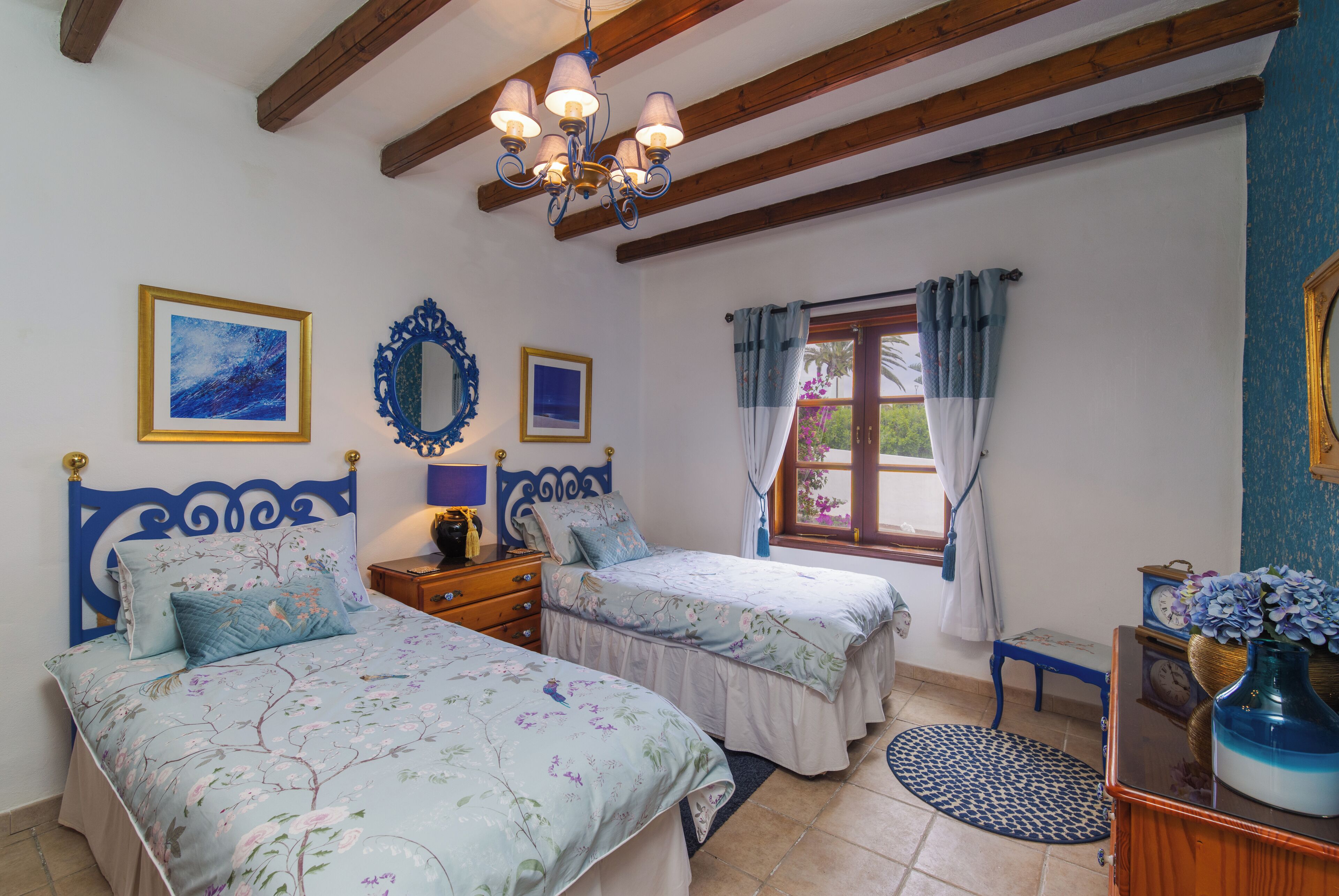 A bedroom with two beds, wooden beams, a chandelier, and a window with curtains.