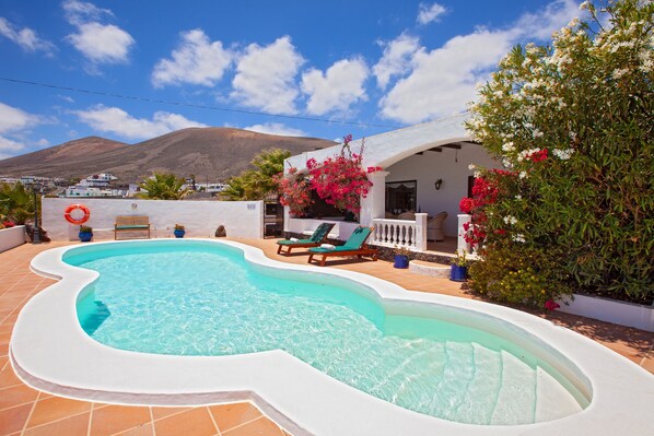 A swimming pool with a wavy edge, surrounded by a white wall and a patio area with two lounge chairs.