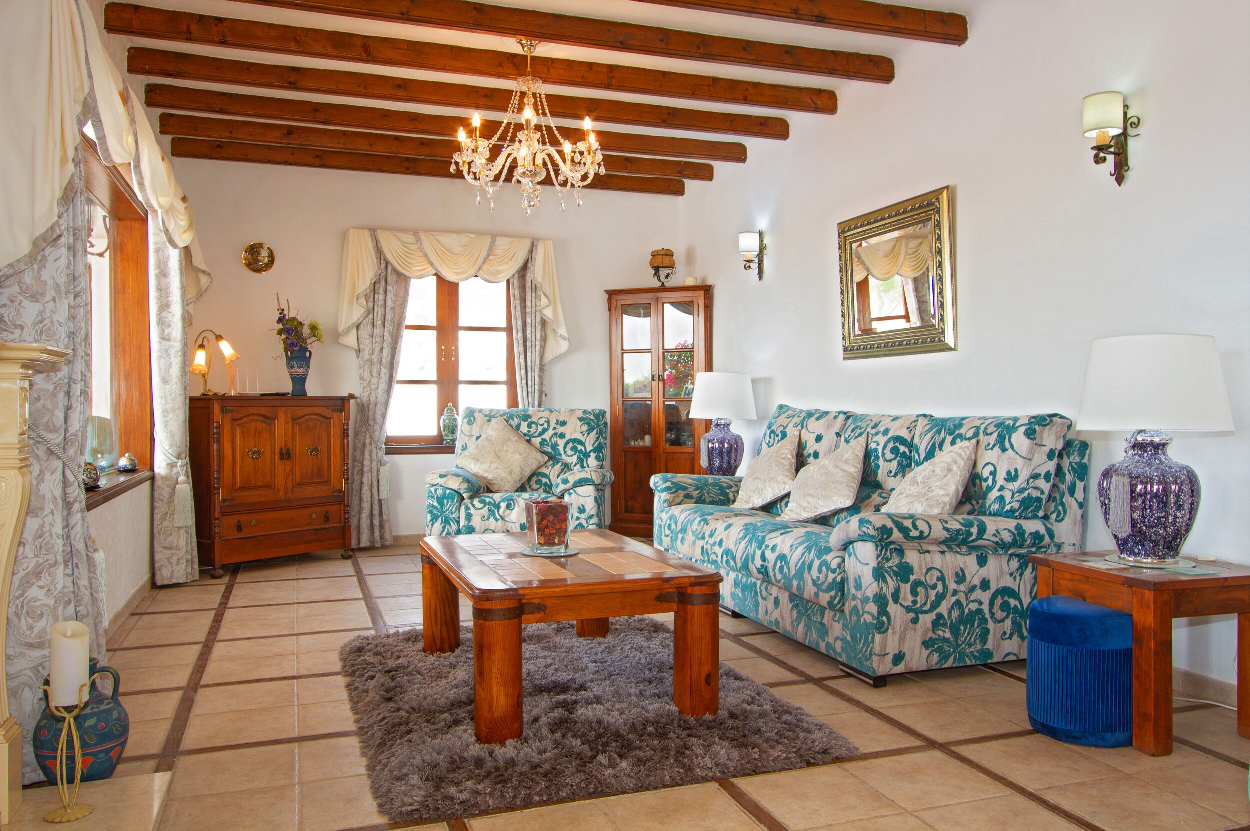 A cozy living room with a wooden ceiling, a chandelier, a sofa with floral upholstery, a wooden coffee table, and a blue ottoman.