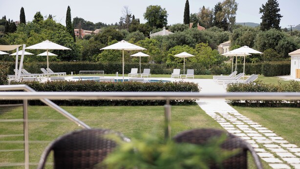 Zona junto a la piscina con sillones de descanso y sombrillas, rodeada de vegetación y un sendero que lleva a un edificio.