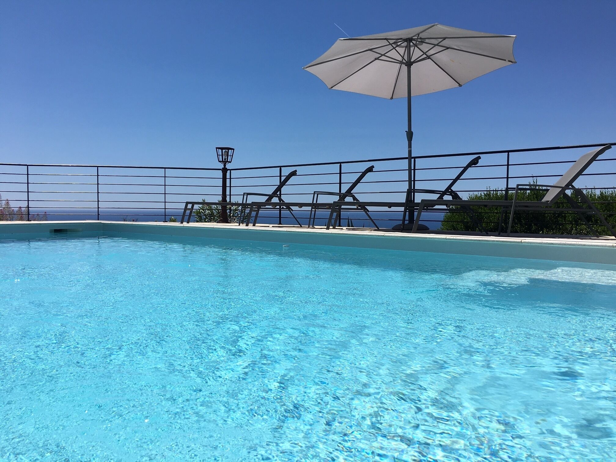 A clear blue pool with a metal fence, a large umbrella, and a view of the ocean.