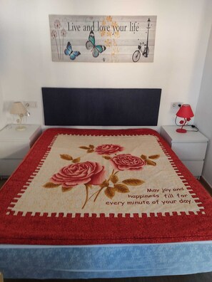 A bedroom with a bed covered in a floral blanket, a nightstand with a lamp, and a wall decoration above the bed. - Torrevieja, 360 days of sunshine to enjoy; spacious apartment, swimming pools and sea, Nl. (Torrevieja)