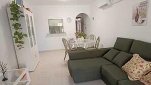 Una sala de estar con un sofá verde, una mesa de comedor con sillas, un ventilador de techo y un mueble blanco con plantas.