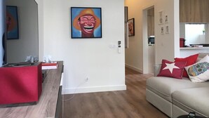 Una sala de estar moderna con un sofá rojo, un sofá blanco y una zona de comedor con una mesa roja. Hay pinturas en las paredes y un equipo de aire acondicionado sobre la zona de comedor.
