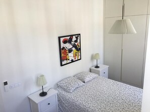 Un dormitorio con una cama, dos mesitas de noche, una lámpara en cada mesita, una obra de arte enmarcada en la pared y un armario.