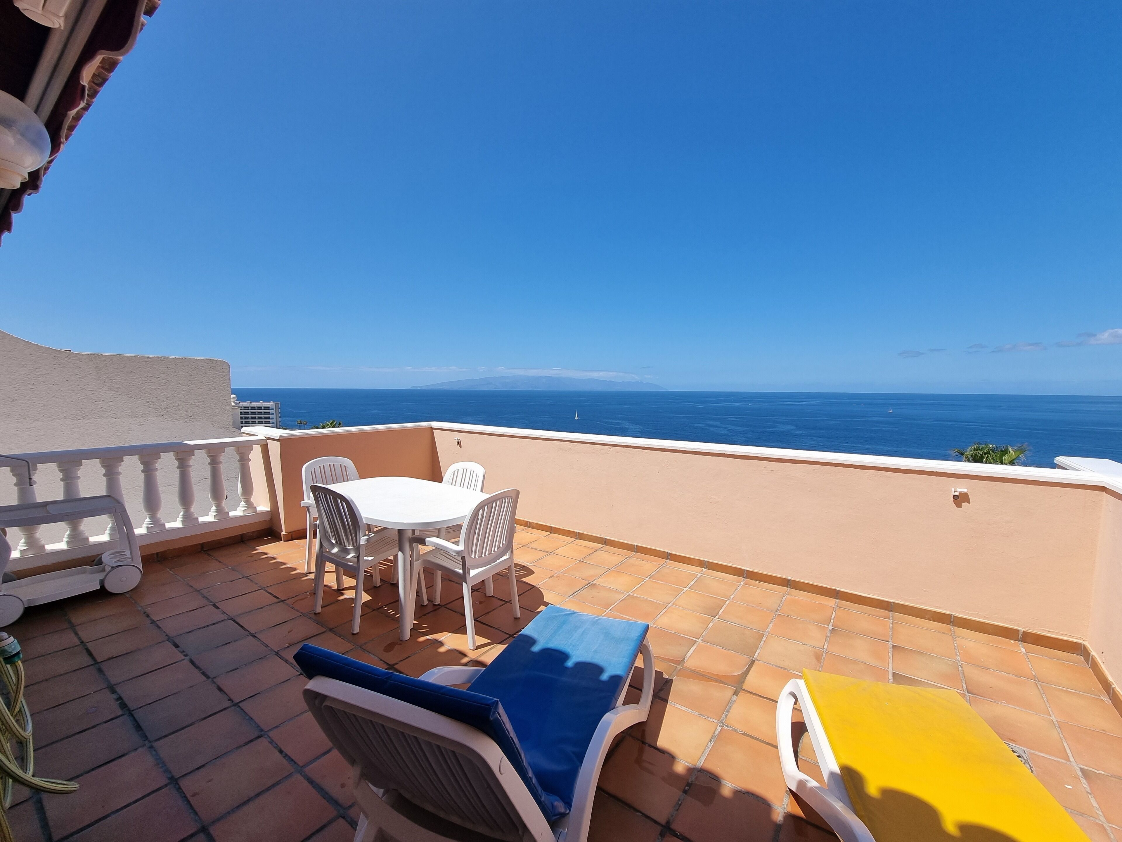 Una terraza con mesa y sillas, una butaca azul y amarilla, y vista al mar.