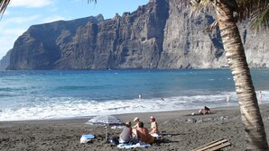A beach with a rocky cliff in the background, a palm tree, and people relaxing on the sand. - APARTMENT WITH SEA VIEWS AND FANTASTIC SUNSHINE (Santiago del Teide)