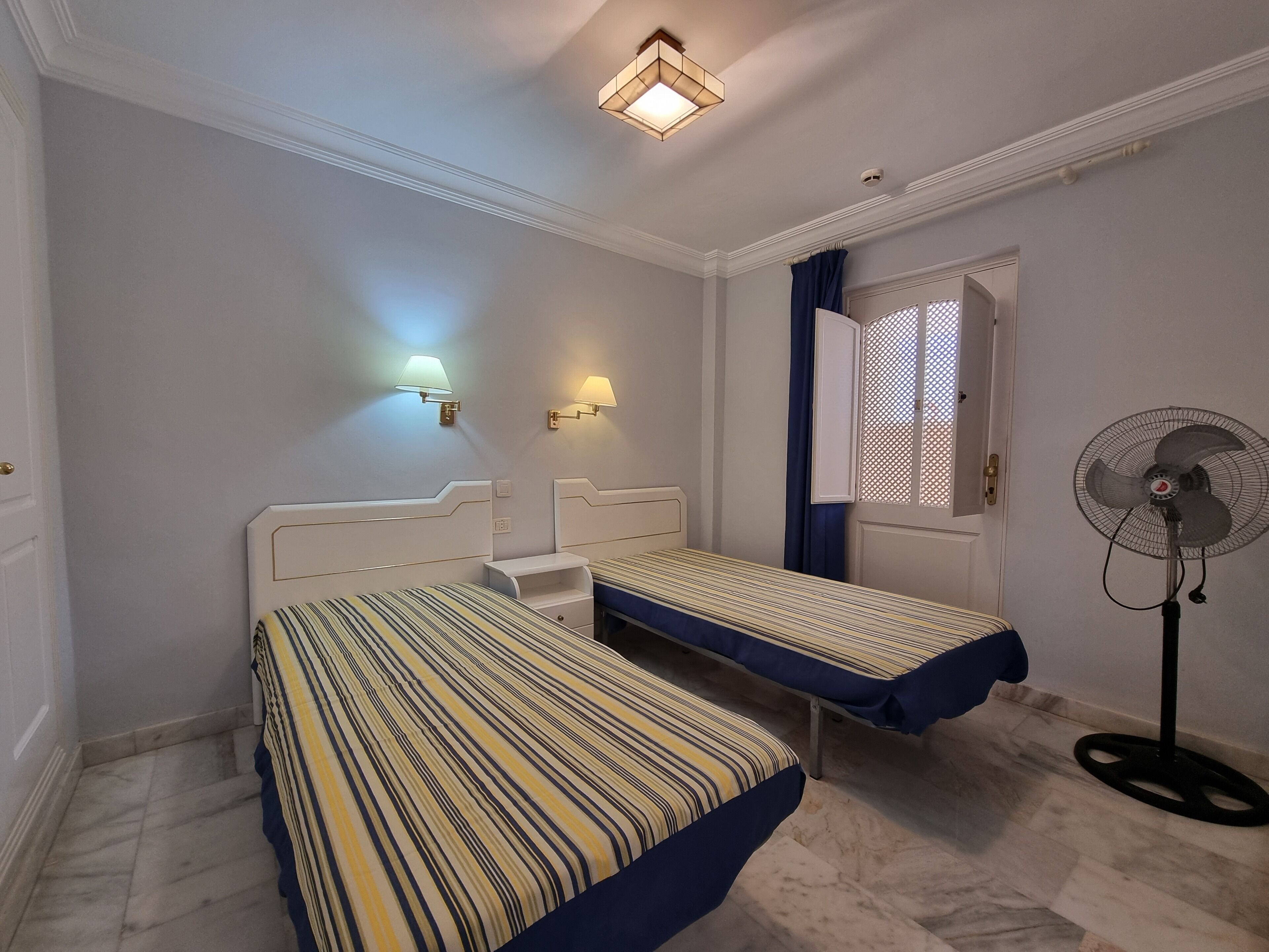 Une chambre d’hôtel avec deux lits simples, un ventilateur sur pied et deux lampes fixées au mur.