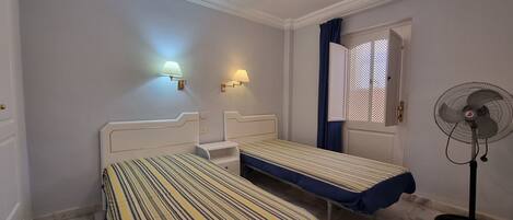 Une chambre d’hôtel avec deux lits simples, un ventilateur sur pied et deux lampes fixées au mur.