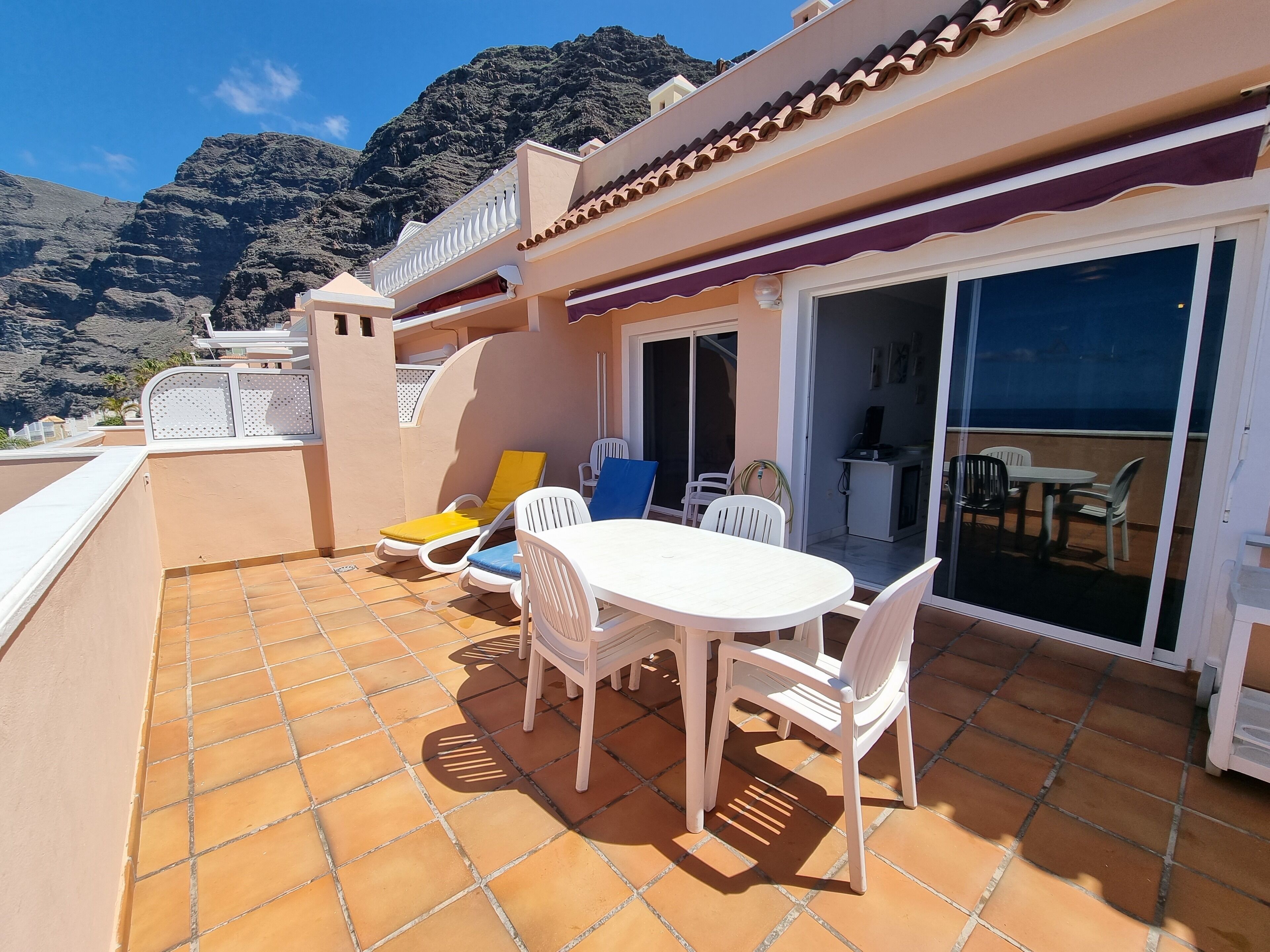 A patio with a white table and chairs, a yellow and blue lounge chair, and a view of mountains in the background.