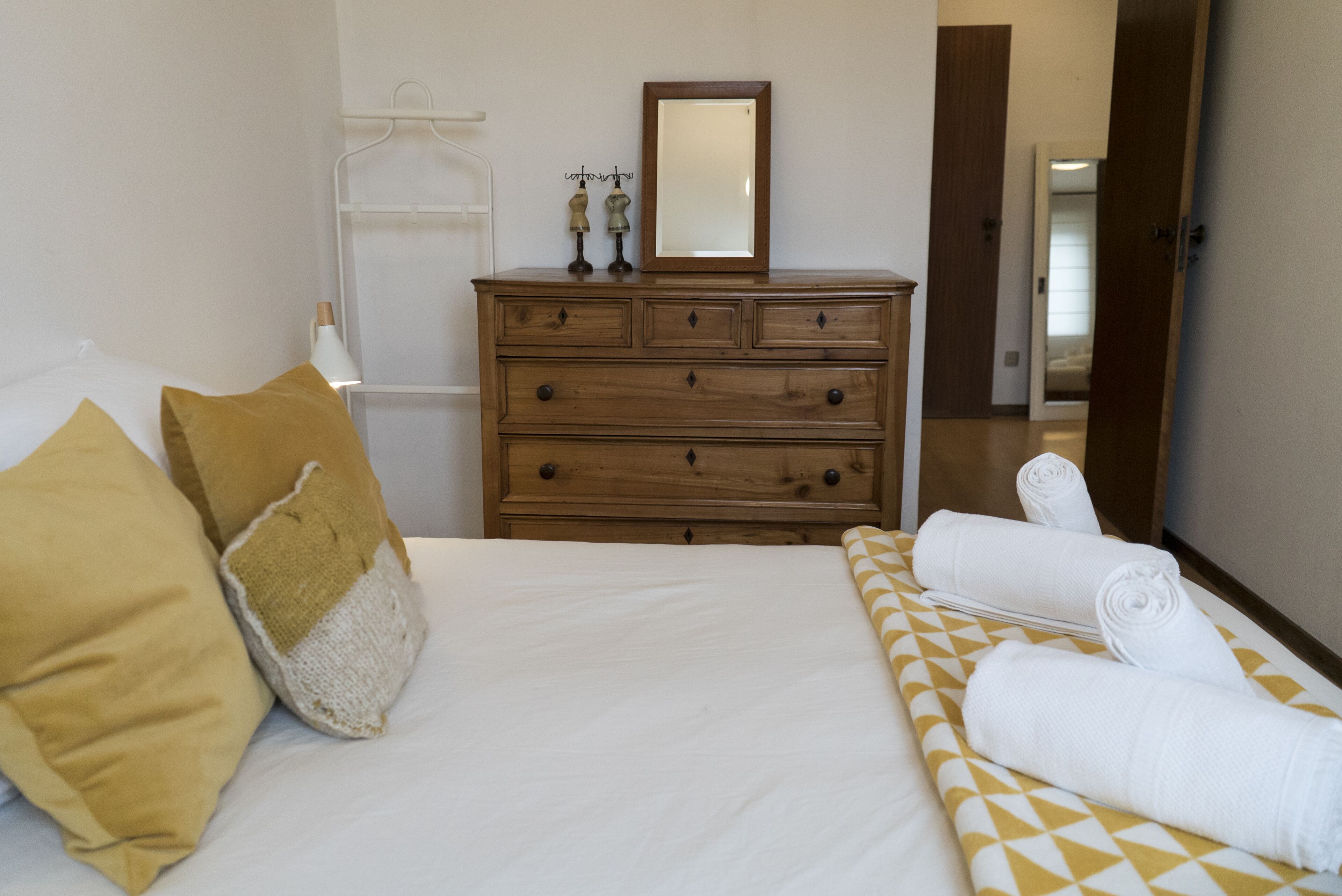 Een netjes opgemaakt bed met witte lakens, een houten dressoir met laden en een spiegel erboven.