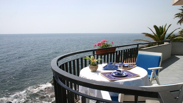 Una mesa redonda con mantel blanco, sillas azules y una planta en maceta con flores rosadas en un balcón con vista al mar.