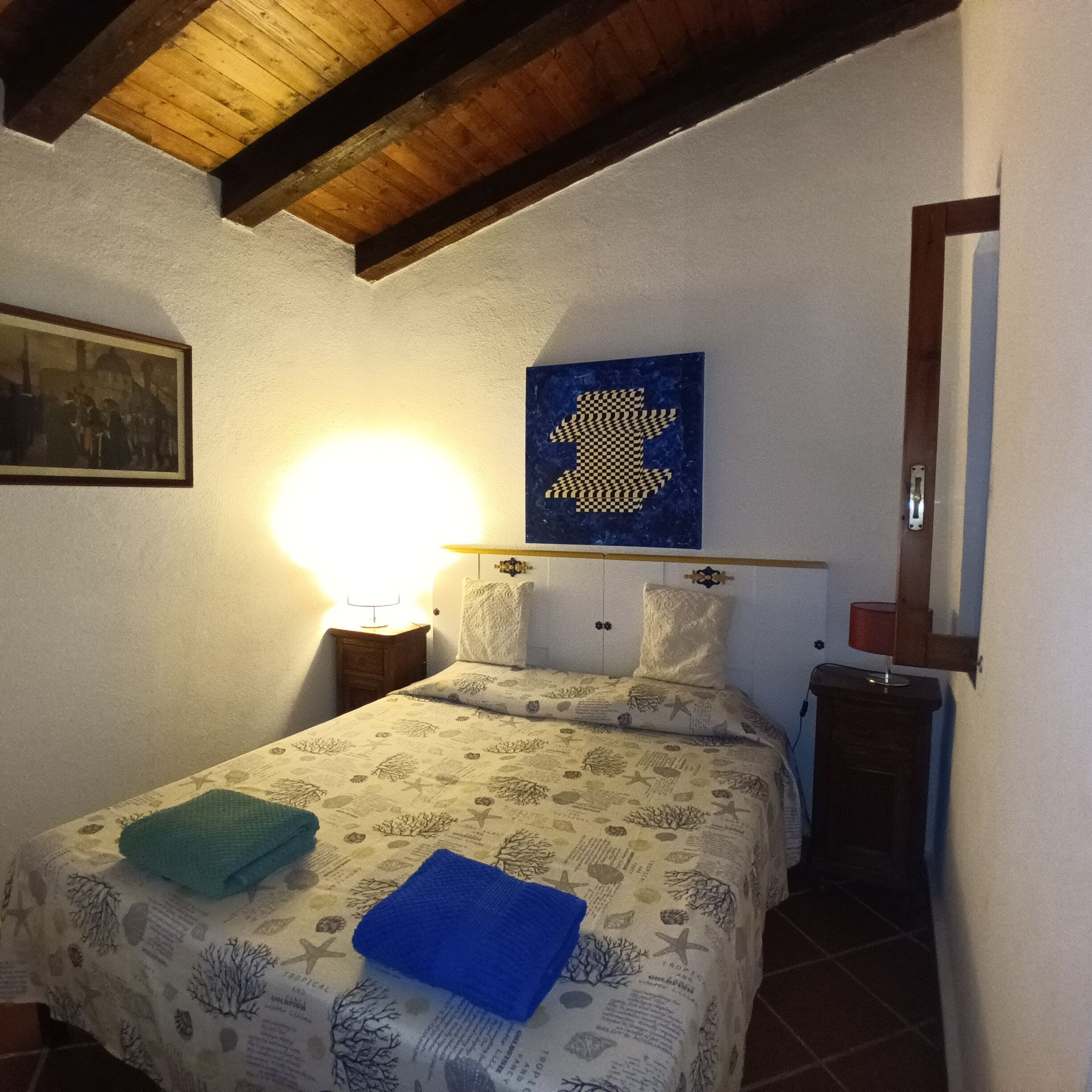 Un dormitorio con cama, mesita de noche, lámpara, cuadro enmarcado y espejo instalado en la pared.