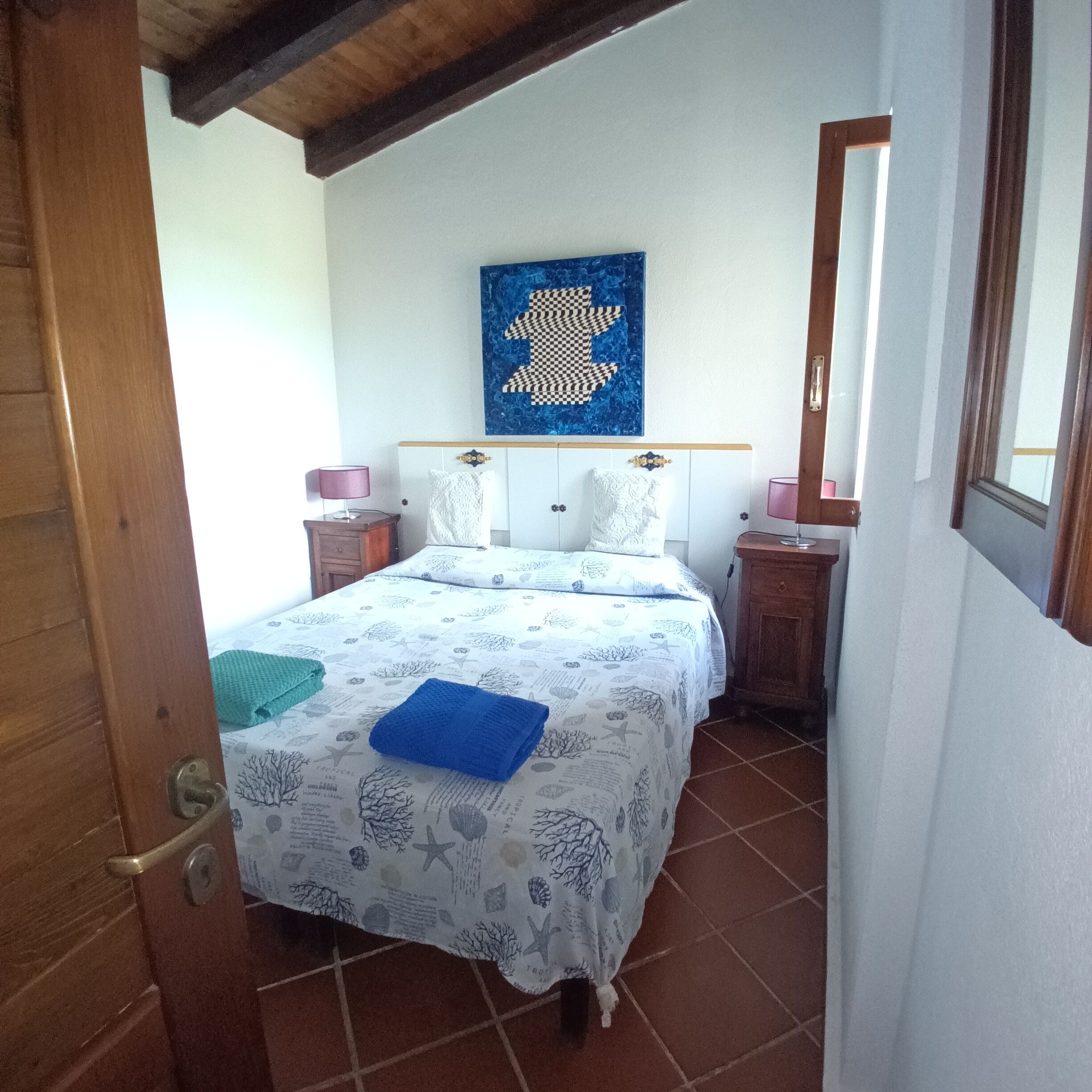 A bedroom with a bed, a nightstand, a dresser, a window, and a wall with a blue artwork.