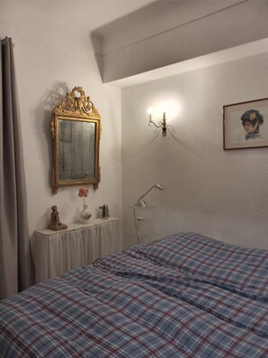 Une chambre à coucher comprenant un lit, un miroir, une lampe fixée au mur, un tableau encadré et une petite table sur laquelle se trouvent un vase et une statue.