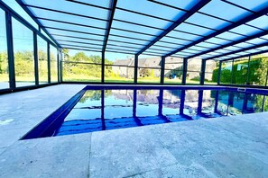 Ein Schwimmbad mit Glasveranda, umgeben von einer Steindeck und großen Fenstern mit Blick auf Grünflächen und Gebäude.