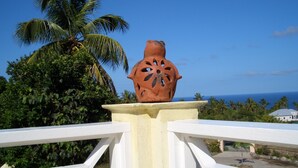 A terracotta pot on a white pedestal with a railing, palm trees, and a clear blue sky in the background. - Villa with large pool, screened terrace overlooking Caribbean. (Jessops)
