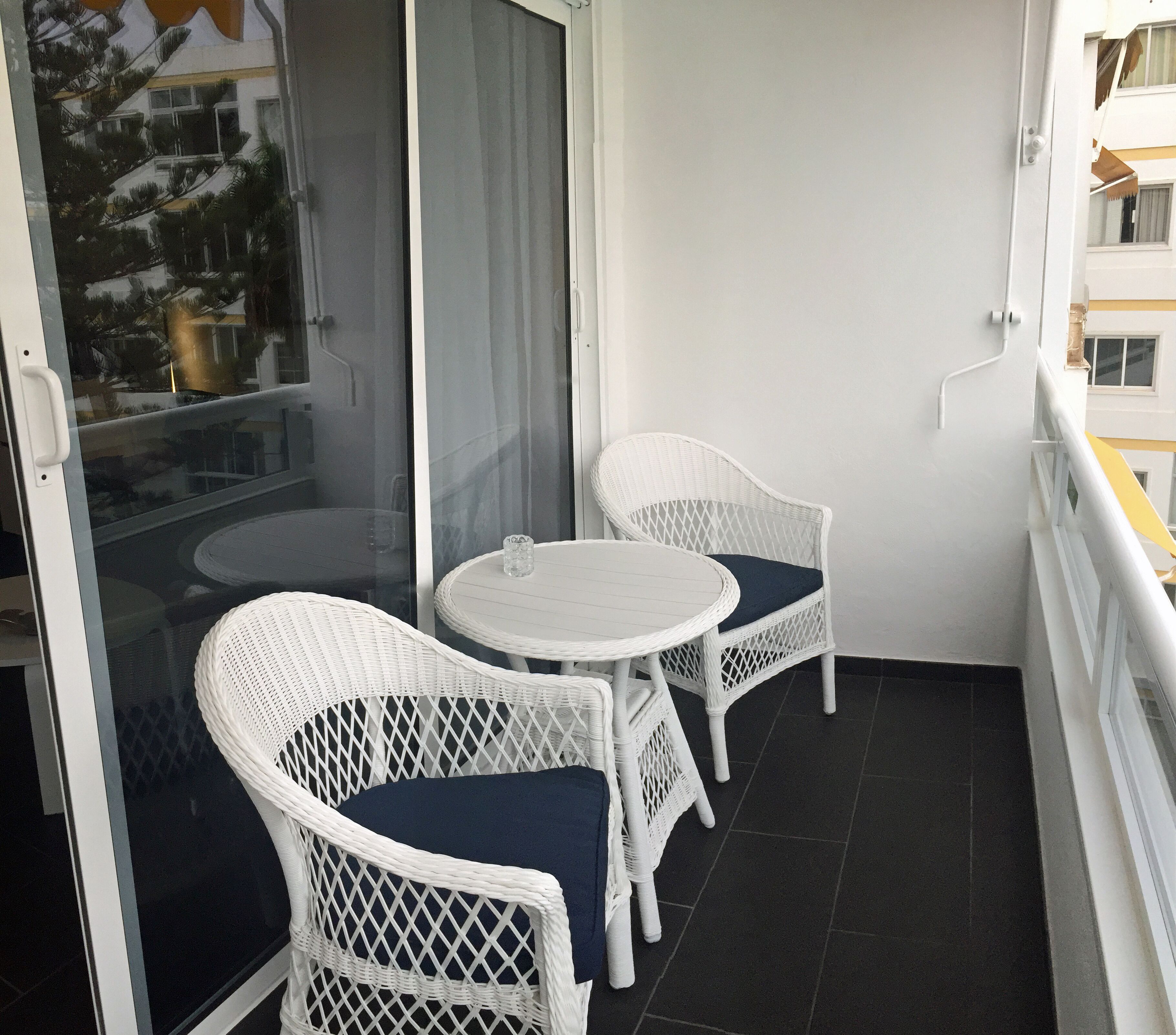 Un balcon agrémenté de deux fauteuils en osier blanc et d’une petite table ronde, avec des coussins aux couleurs sombres.