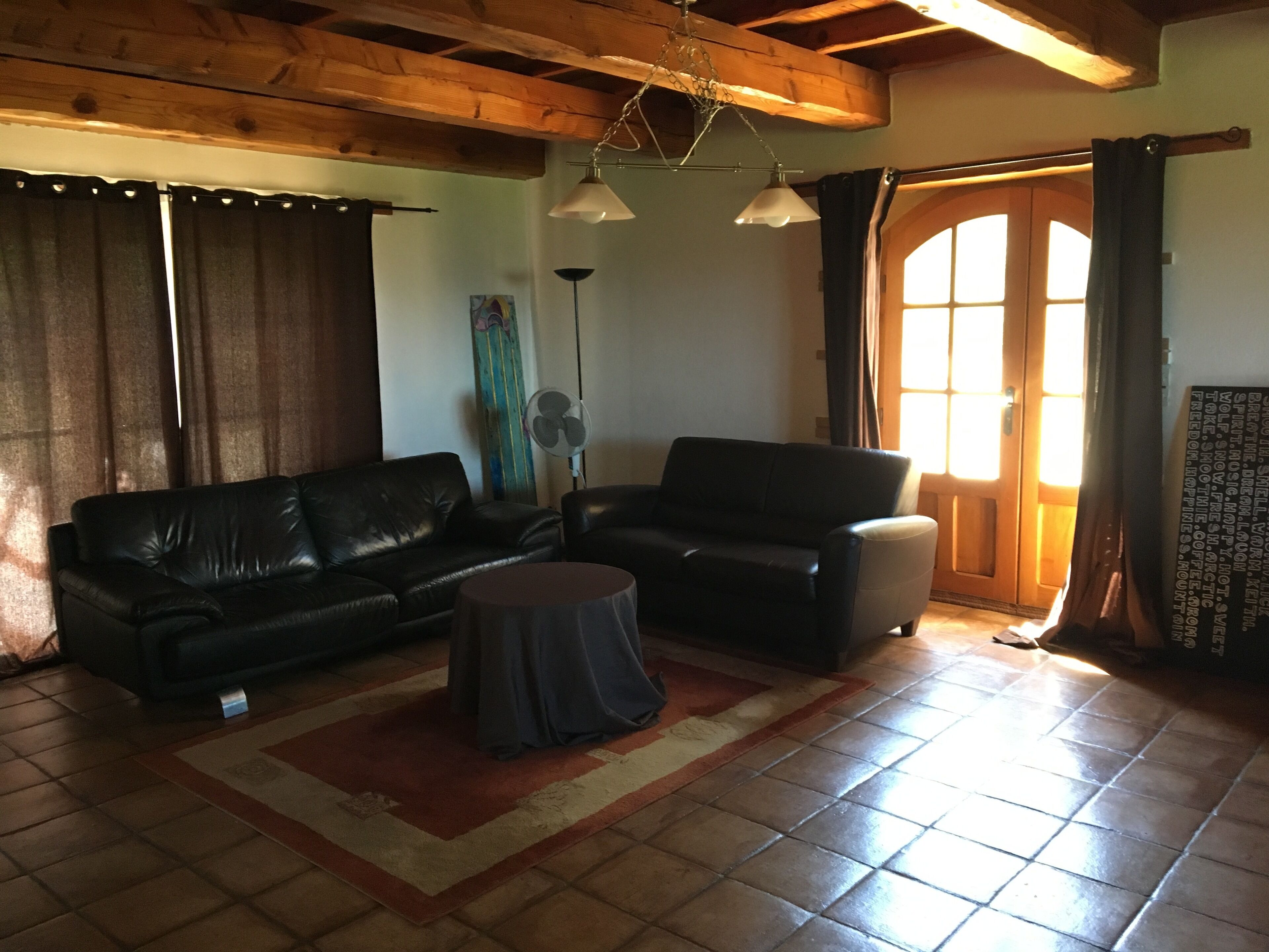 Sala de estar com sofá de couro preto, mesa de centro redonda e um ventilador. Teto de madeira e porta com cortinas.